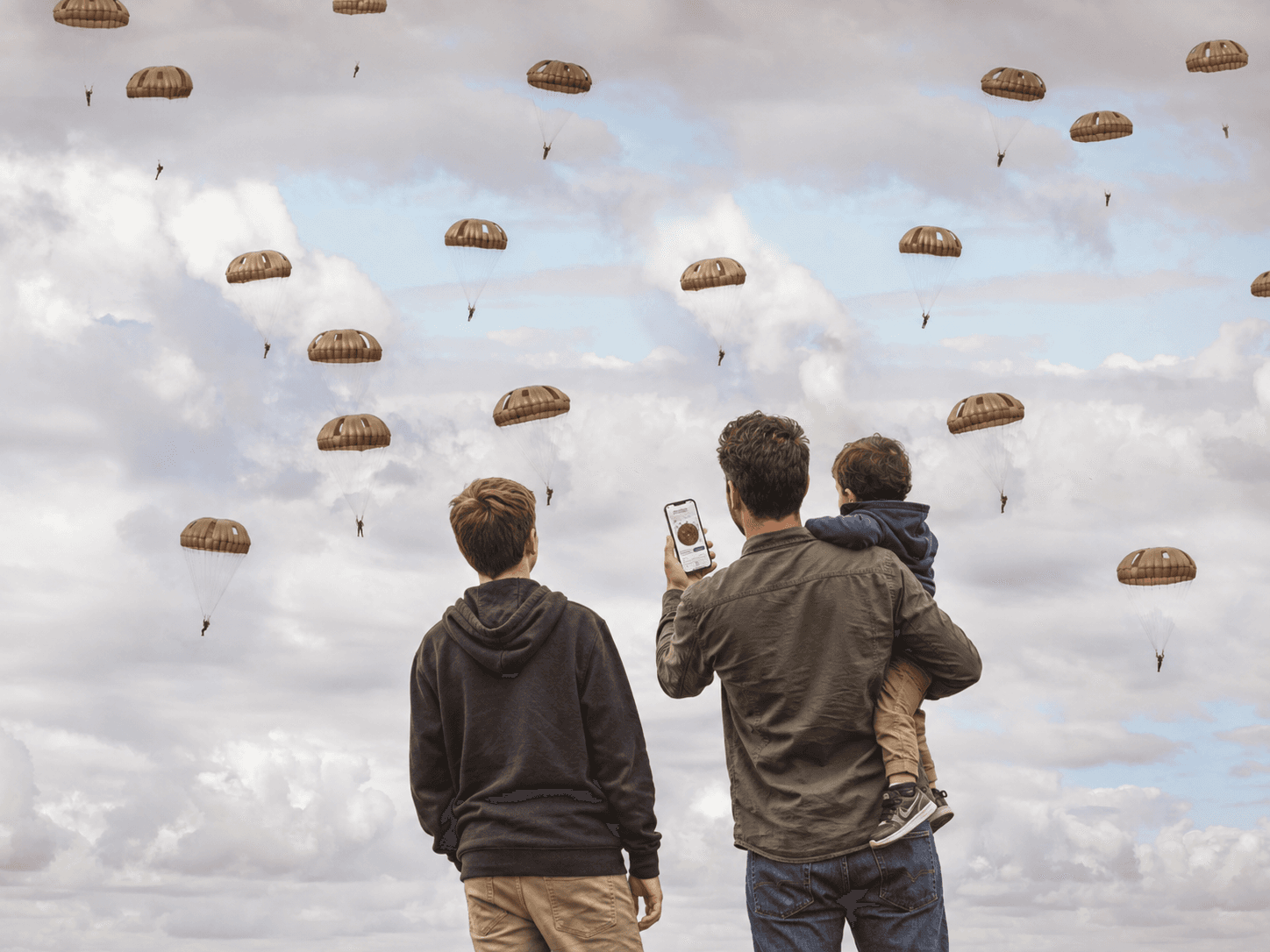 A parent and two children watching a mass parachute drop, phone in hand, with the Ginkelse Heide souvenir coin they can claim there